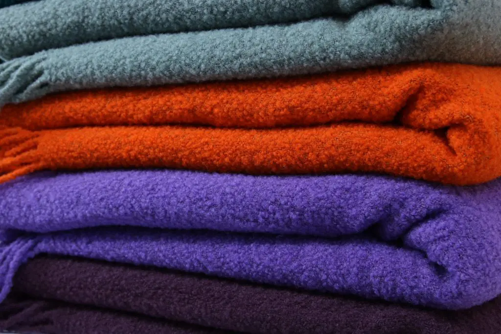 Die gemütlichen Wolldecken von Aufschnitt aus Berlin liegen zusammengelegt übereinander. Man kann die Farben rot, lila, Aubergine und Salbei sehr gut erkennen und sich vorstellen, wie weich sie sich anfühlen.
